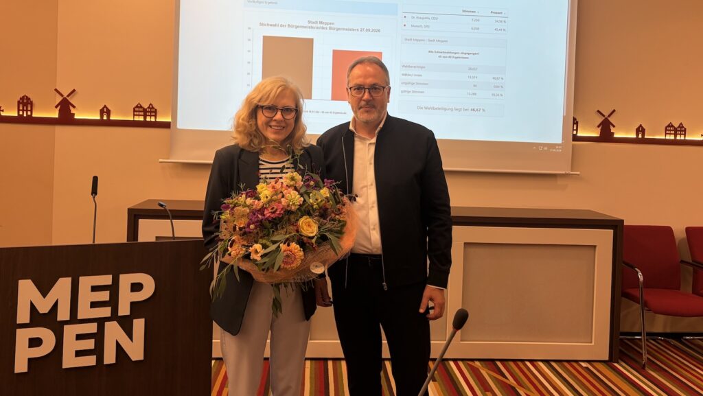 Dr. Sigrid Kraujuttis mit Blumenstrauß neben einem Gratulanten im Ratssaal Meppen, im Hintergrund die Präsentation der Wahlergebnisse