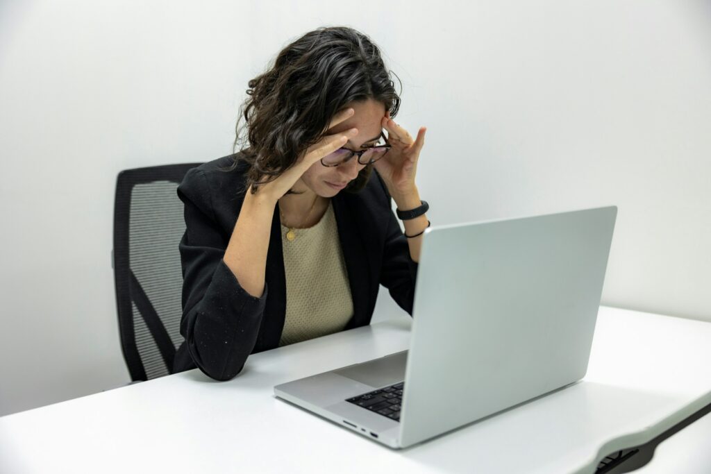 Erschöpfte Mitarbeiterin fasst sich mit schmerzverzerrtem Gesicht an den Kopf, im Hintergrund ihr Laptop am Schreibtisch