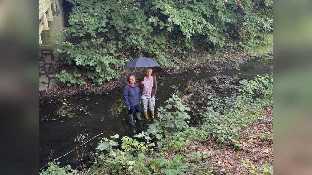 Zwei Personen mit Regenschirm stehen im Wasser eines Baches am Ufer