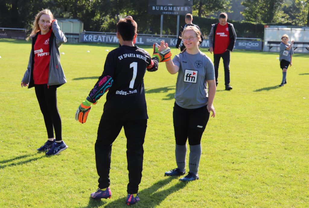 Mannschaften und Zuschauer stehen sich auf dem Sportplatz gegenüber, Spieler heben die Hand