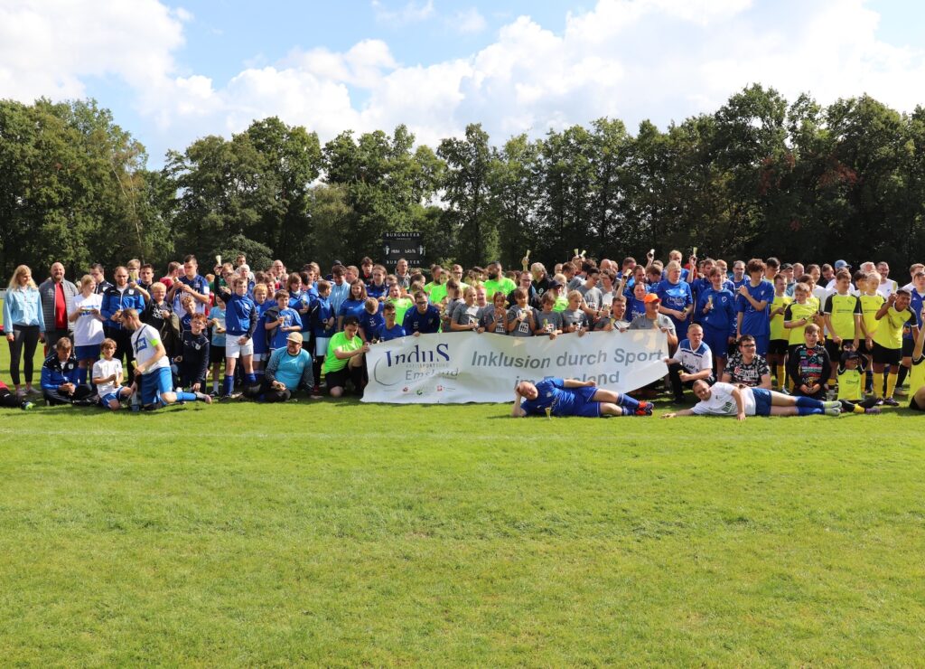 Ein Torwart begrüßt einen jungen Spieler auf dem Fußballplatz, im Hintergrund weitere Kinder und Häuser