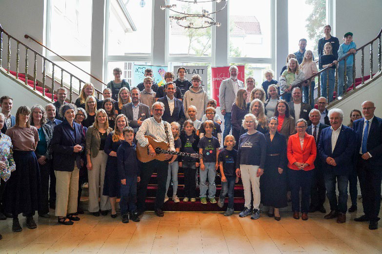 Ministerpräsident Olaf Lies steht mit Gitarre inmitten von Kindern, Jugendlichen und Erwachsenen der Arbeitsgruppe Platt is cool auf einer Treppe in der Niedersächsischen Staatskanzlei