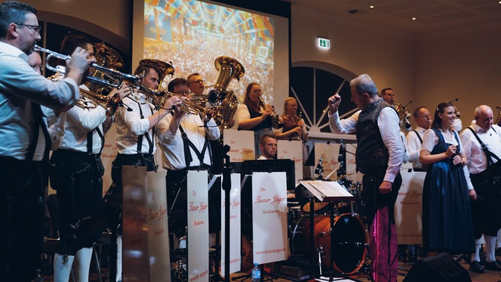 Die Tinner Jäger Musikanten spielen auf der Bühne, im Hintergrund ist auf einer Leinwand ein Festzelt zu sehen