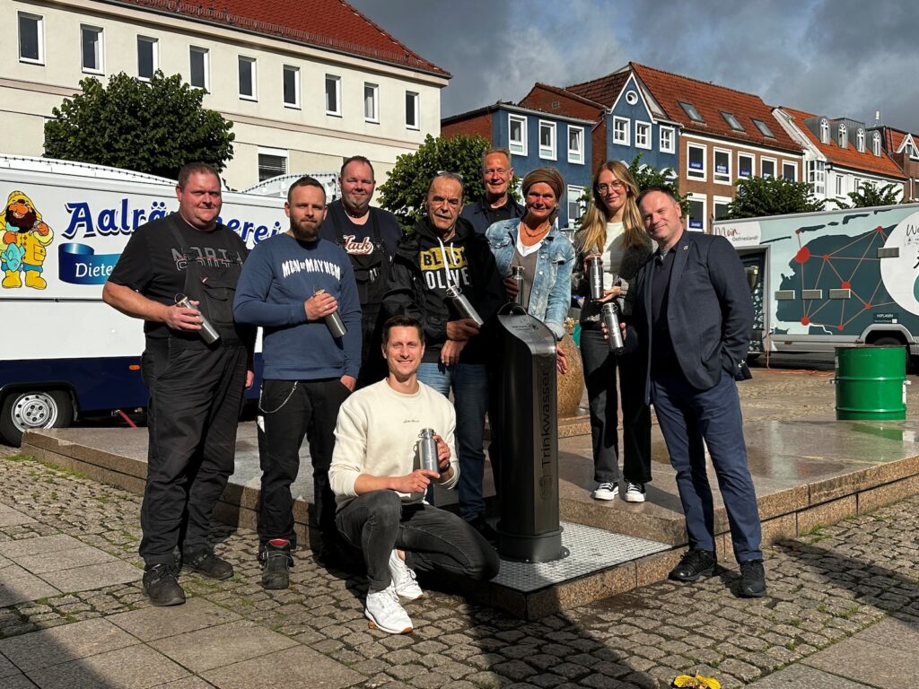 Mitarbeiter der Stadt Aurich und beteiligte Institutionen stehen bei der Einweihung um den neuen Trinkwasserbrunnen auf dem Auricher Marktplatz