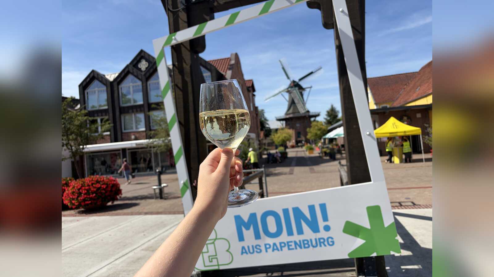 Weinglas vor einem MOIN-AUS-PAPENBURG-Rahmen mit Blick auf die Papenburger Altstadt und eine Windmühle