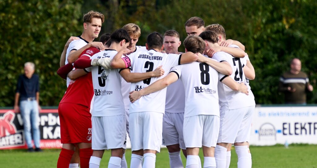 Spieler des SC Spelle-Venhaus in weißen Trikots stehen beim Derby in Hemsen im Kreis zusammen