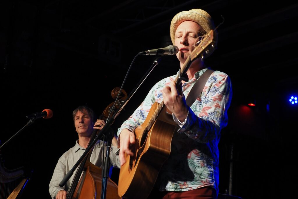 Toni Geiling mit Strohhut singt und spielt Gitarre, dahinter ein Kontrabassist auf der Bühne