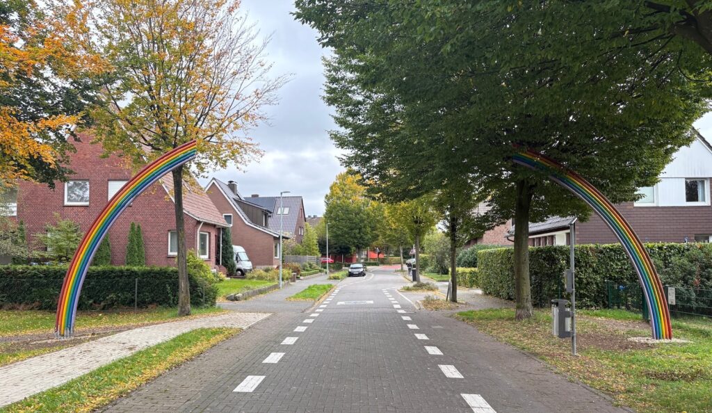 Ein bunter Regenbogenbogen aus Metall überspannt eine gepflasterte Fahrradstraße zwischen Bäumen und Wohnhäusern