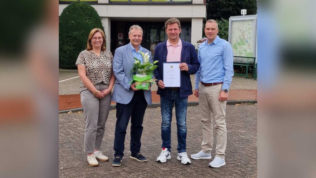Vier Personen vor dem Holdorfer Rathaus, Thomas Oeverhaus hält eine Urkunde und einen Blumentopf