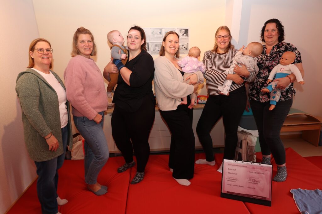 Mütter mit ihren Babys und Kursleiterinnen stehen in einem Kursraum mit roten Matten