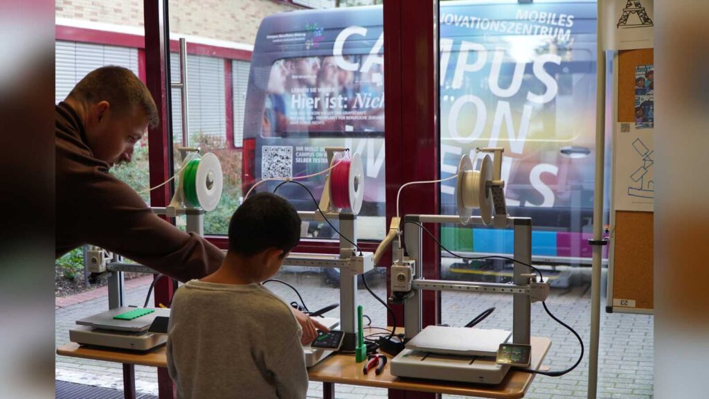 Ein Junge arbeitet mit einem Betreuer an 3D-Druckern, im Hintergrund der Transporter Campus on Wheels
