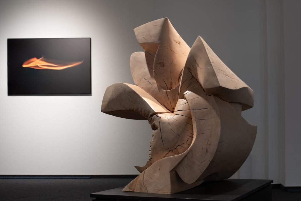 Große Holzskulptur mit geschwungenen Formen vor einer Wand mit einem abstrakten Lichtbild in Orange auf Schwarz