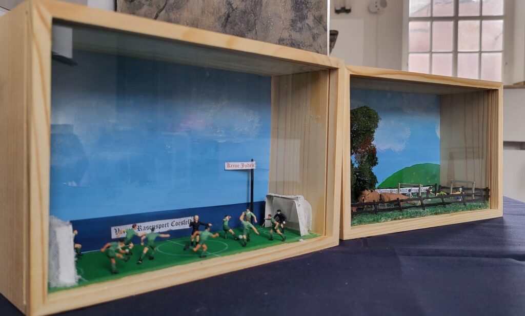 Zwei Schaukästen mit Dioramen, links ein Fußballspiel mit einem Schild Keine Juden, rechts eine Weide mit Kindern hinter einem Zaun