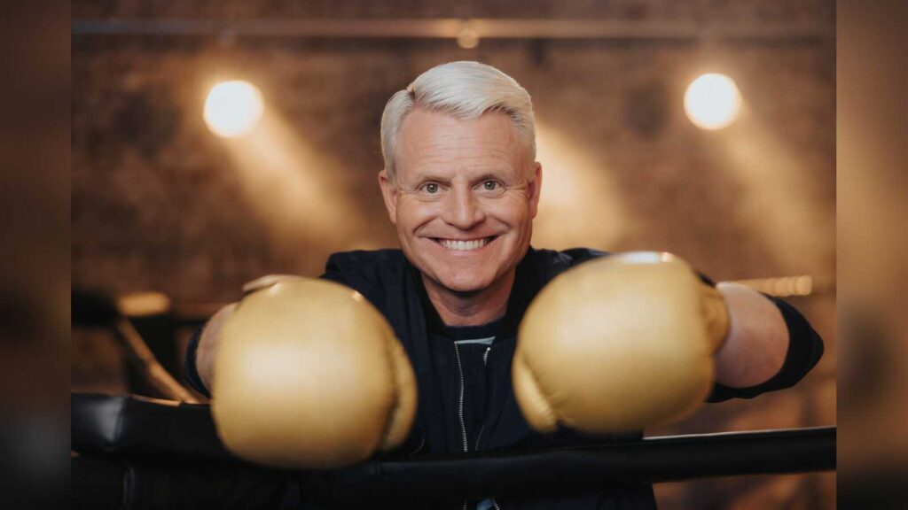 Guido Cantz lehnt lachend mit goldenen Boxhandschuhen über den Seilen eines Boxrings