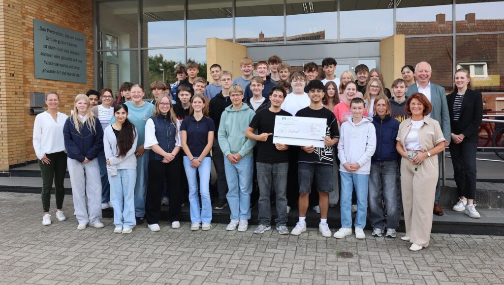 Schülerinnen und Schüler der Klassen 10a und 10b der Georg-Kerschensteiner-Schule Holdorf mit ihren Klassenlehrerinnen und Vertretern der Volksbank Dammer Berge vor dem Schulgebäude, in der Mitte ein Scheck über 2.000 Euro