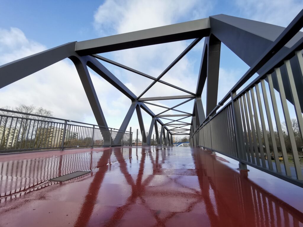 Blick über eine stählerne Fachwerkbrücke für Fußgänger und Radfahrer mit rotem Belag