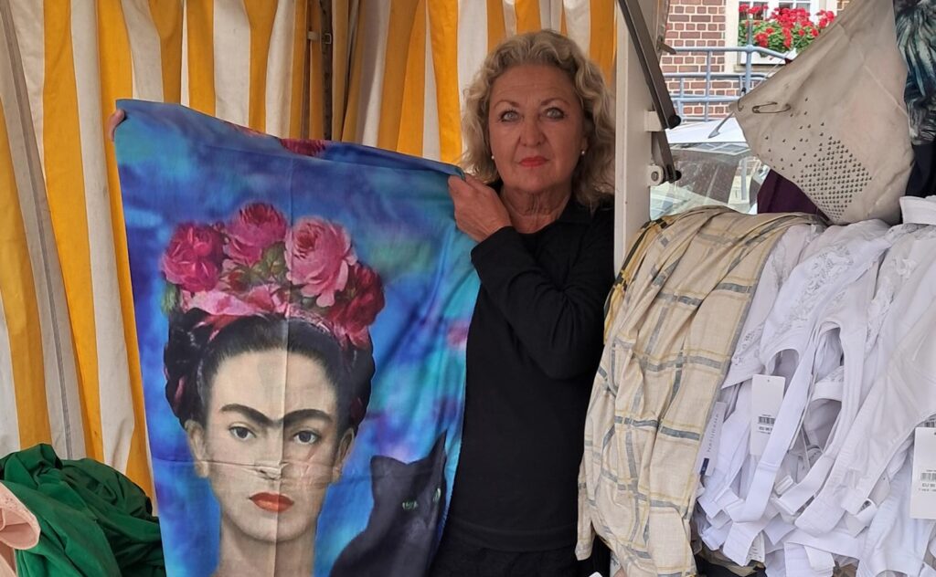 Händlerin Christa Endrigkeit hält an ihrem Marktstand ein bedrucktes Tuch hoch, davor Wäsche und Miederwaren