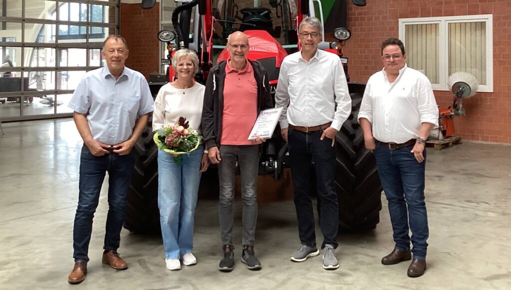 Jubilar Ludger Ahrens mit Urkunde, seine Frau Inge mit Blumenstrauß und drei Gratulanten vor einem roten Traktor in einer Werkhalle in Lastrup