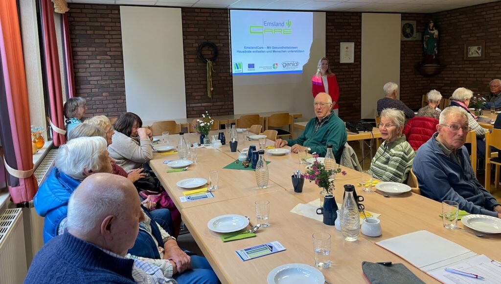 Seniorinnen und Senioren an langen Tischen, vorne steht Franziska Fehnker vor einer Leinwand mit dem EmslandCare-Logo