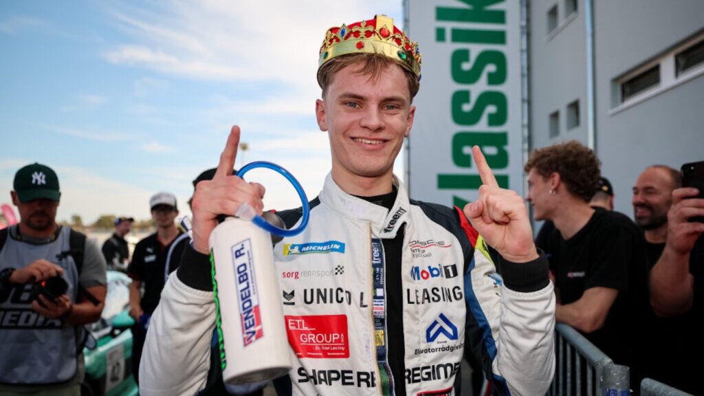 Rennfahrer Rasmus Vendelbo im Rennanzug mit Spielzeugkrone auf dem Kopf zeigt im Fahrerlager des Hockenheimrings zwei Finger in die Höhe