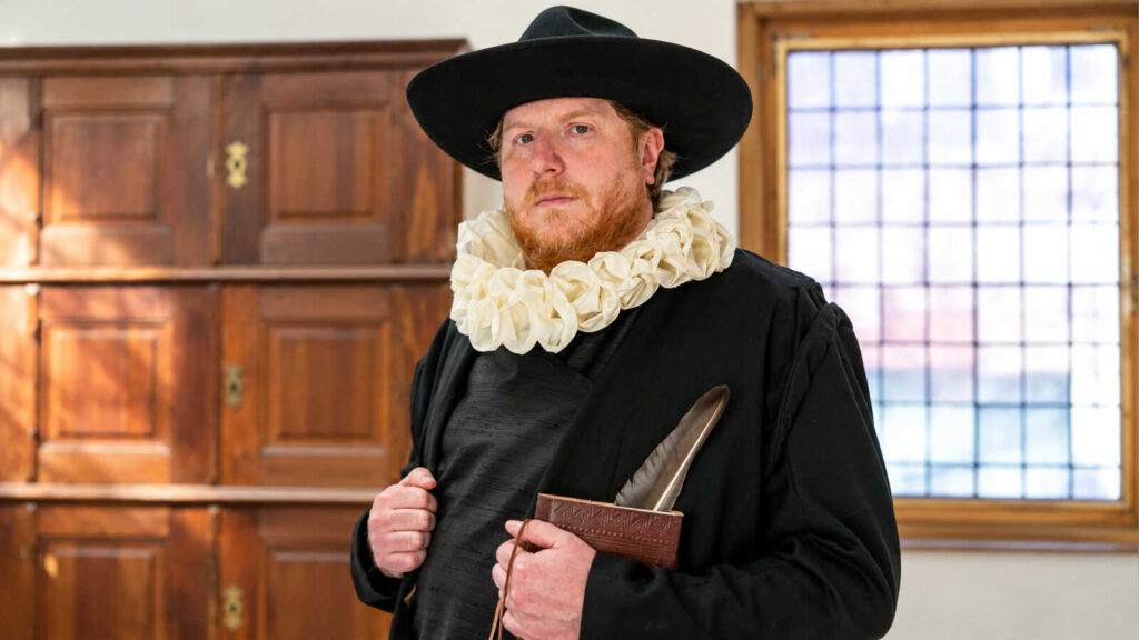 Schauspieler im Kostüm des 17. Jahrhunderts mit schwarzem Hut, weißer Halskrause, Buch und Federkiel in einem Raum von Burg Vischering
