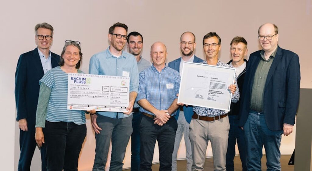 Gruppe von neun Personen mit Spendenscheck über 2.000 Euro und gerahmter Urkunde des Gewässerwettbewerbs Bach im Fluss