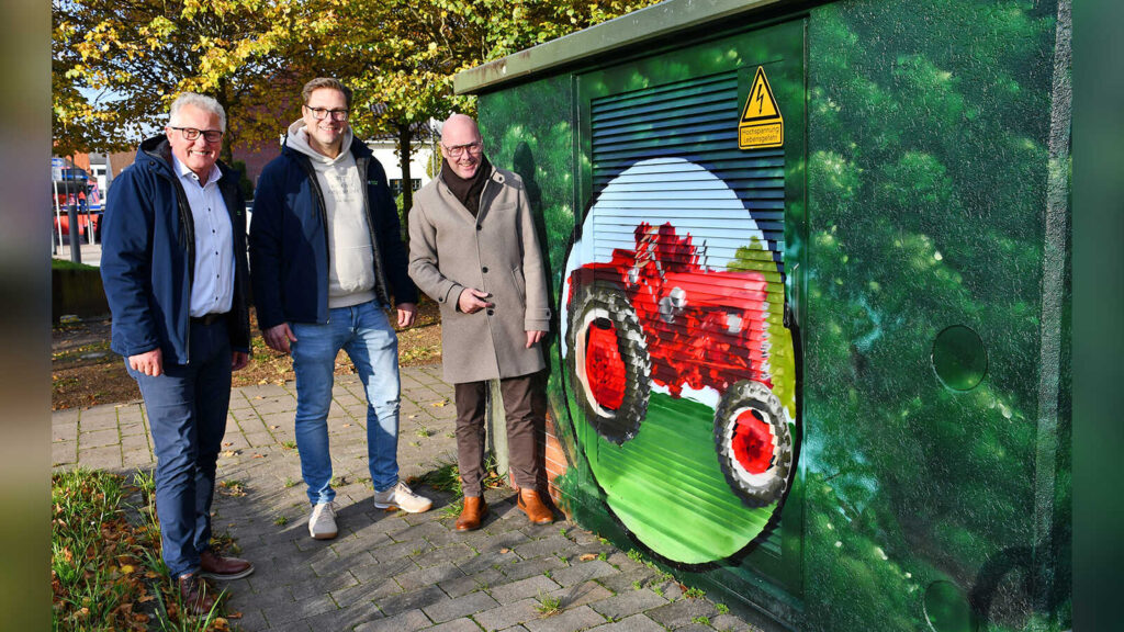 Thomas Lüllmann, Gerd Niemann und Bürgermeister Neidhard Varnhorn vor einer Trafostation mit einem gemalten roten Trecker