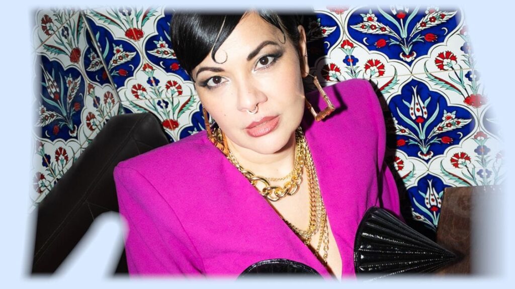 Reyhan Şahin in pinkfarbenem Blazer und Goldketten vor einer Wand mit blau-roten Ornamentfliesen