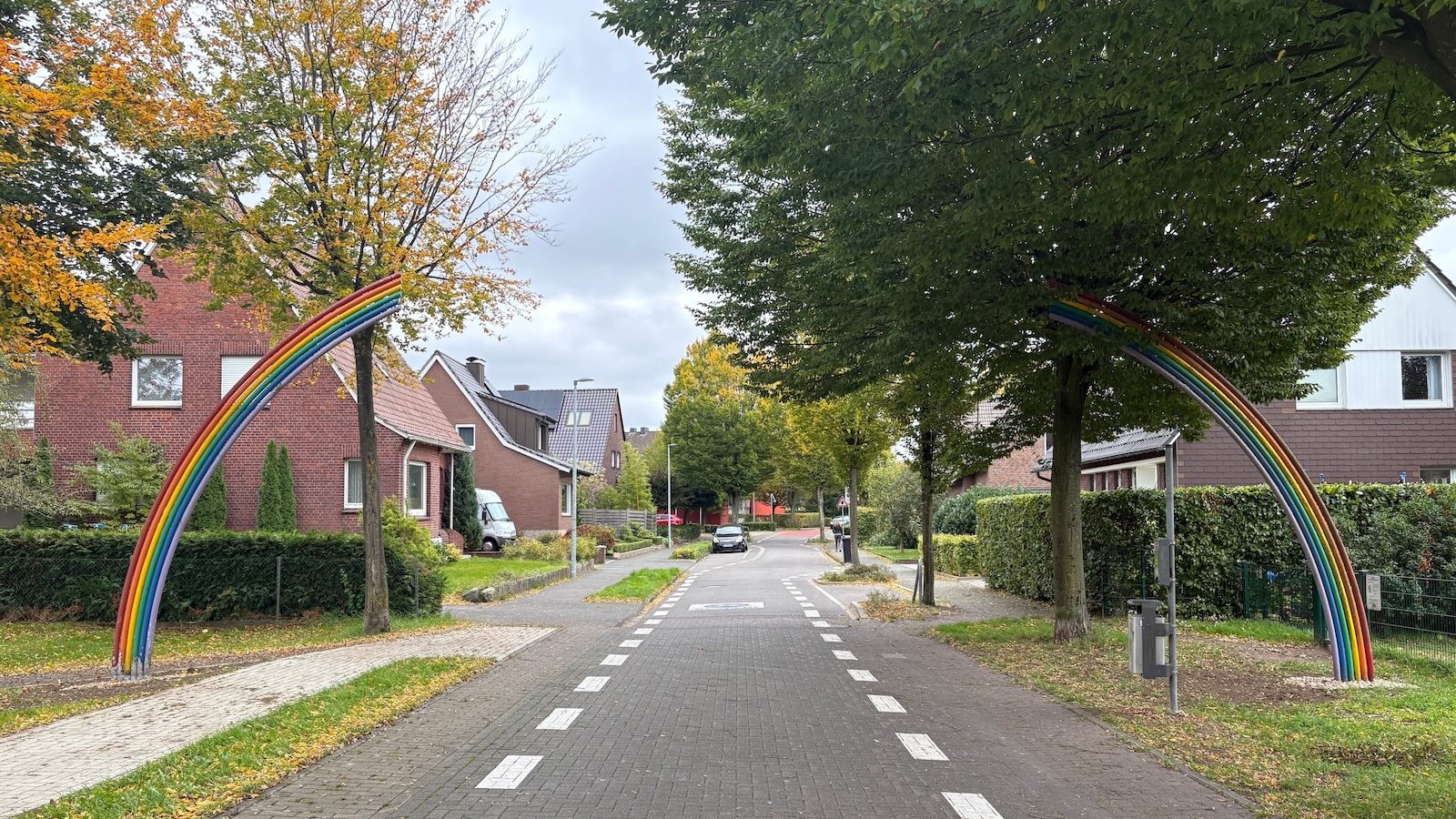 Ein bunter Regenbogenbogen aus Metall überspannt eine gepflasterte Fahrradstraße zwischen Bäumen und Wohnhäusern