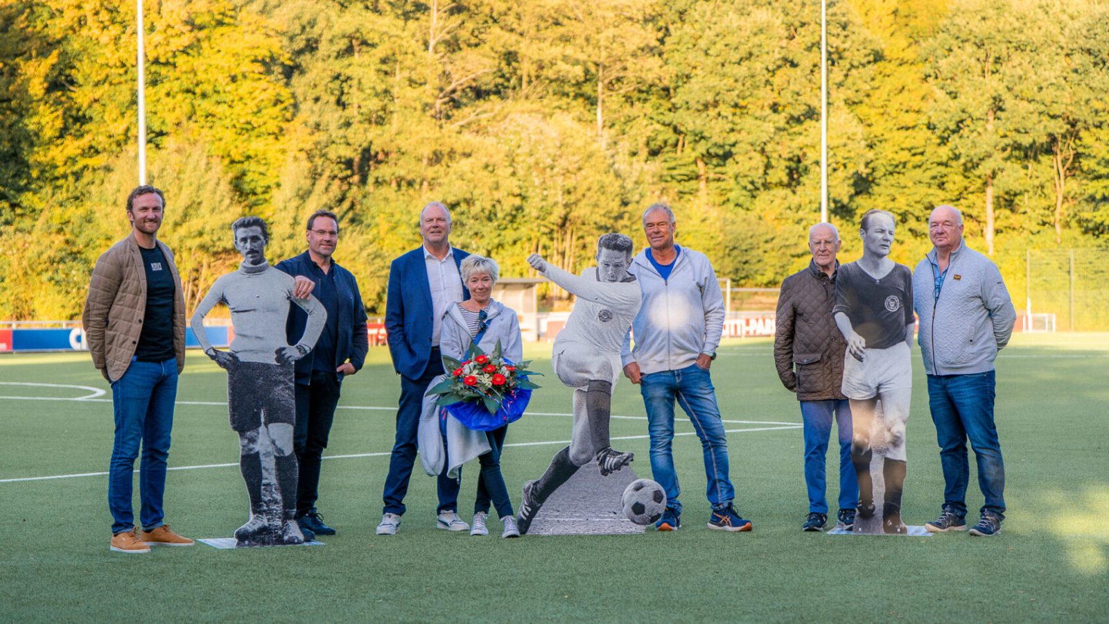Menschen posieren neben Pappfiguren von Fußballspielern auf einem Sportplatz im Freien, eine Person hält Blumen.