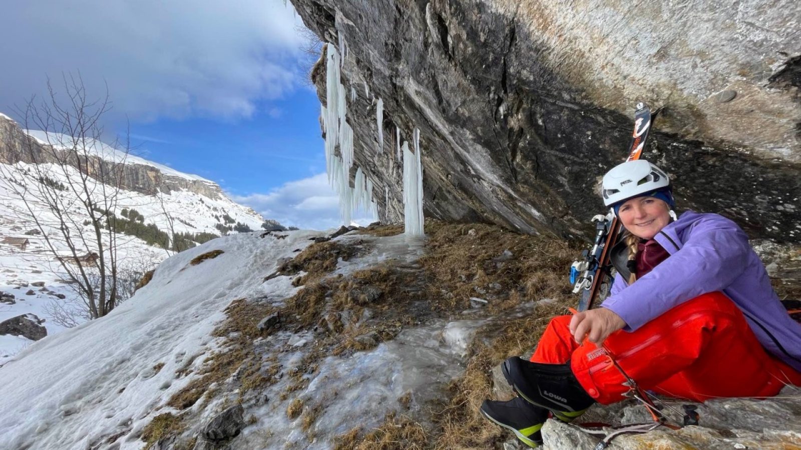 Eine Person in Kletterausrüstung sitzt auf einem Felsen unter einer steilen Felswand mit Eiszapfen.