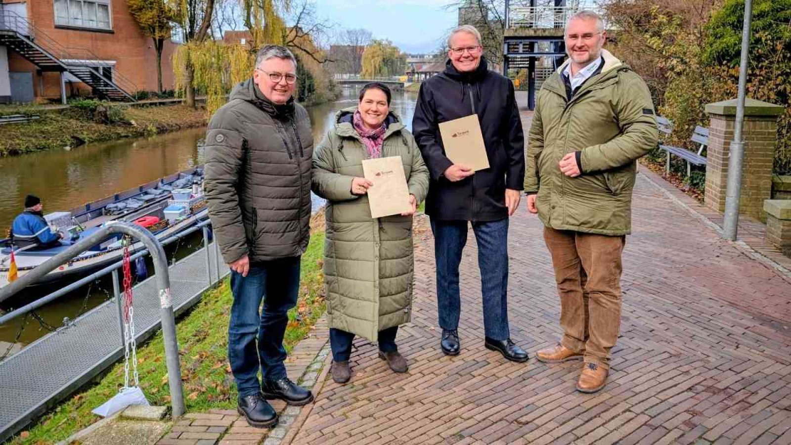 Vier Personen stehen am Ufer eines Kanals und halten Dokumente in den Händen.
