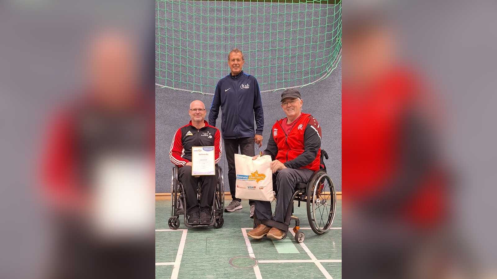 Thomas Hilmes mit Urkunde und Geschenktasche im Rollstuhl, daneben Willi Fenslage und Günter Hömmeke in der Sporthalle