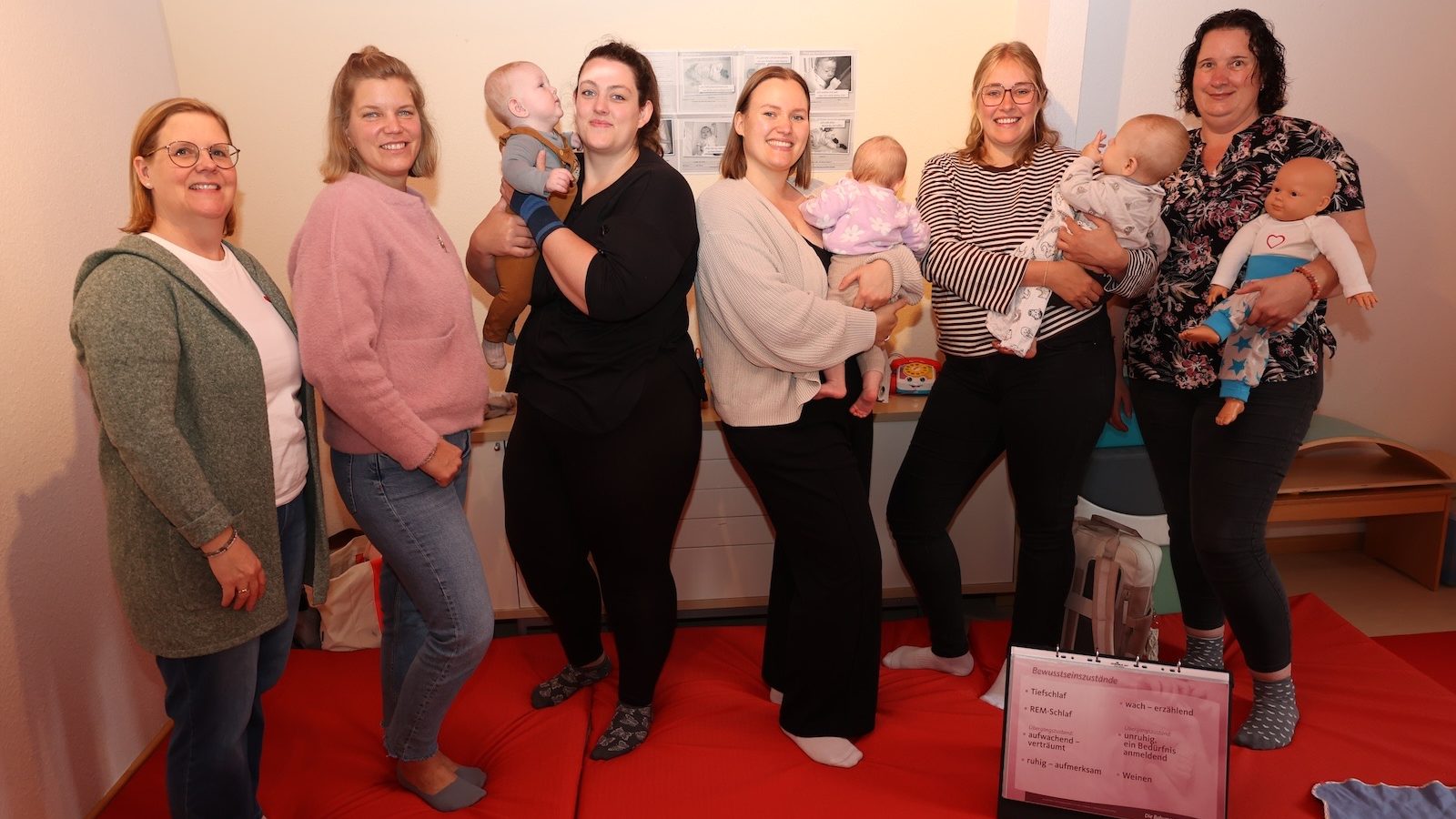 Mütter mit ihren Babys und Kursleiterinnen stehen in einem Kursraum mit roten Matten