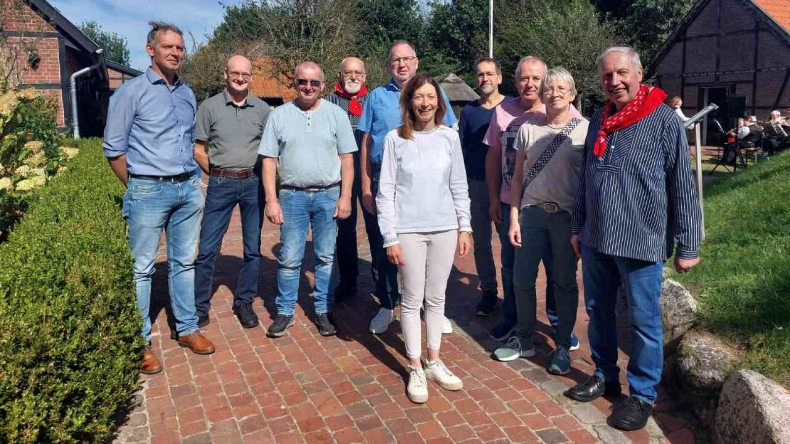 Gruppe von Menschen im Freien an einem sonnigen Tag, auf einem Pflasterweg bei einem Backsteinhaus.