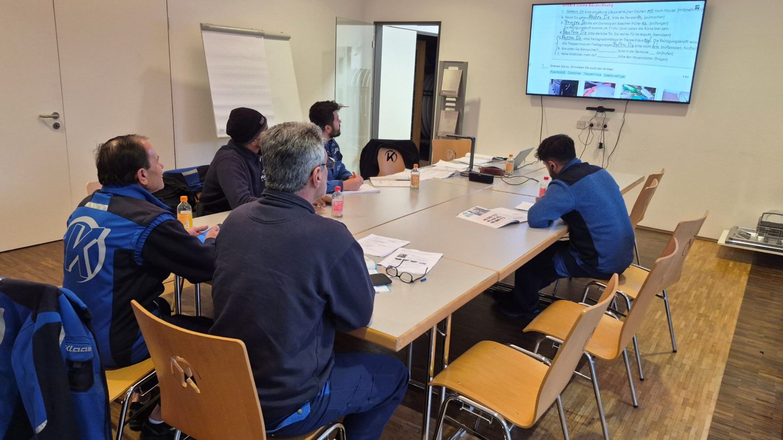 Gruppe von fünf Personen in einem Besprechungsraum, die an einem Tisch sitzen und auf einen Bildschirm schauen.