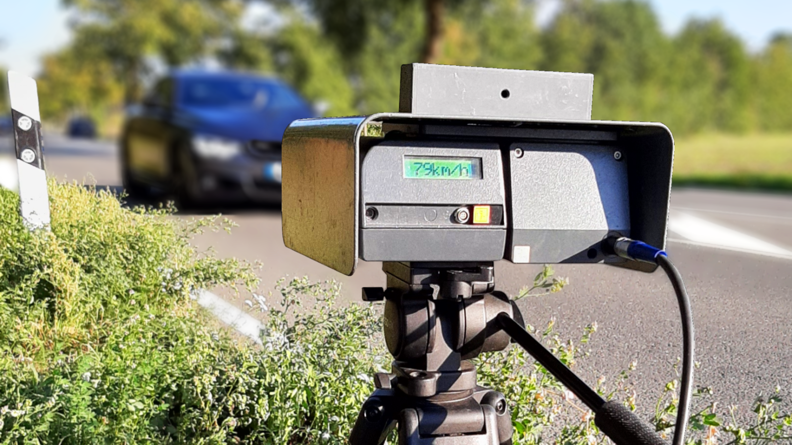 Geschwindigkeitsmessgerät auf einem Stativ an einer Straße, mit einem Fahrzeug im Hintergrund.