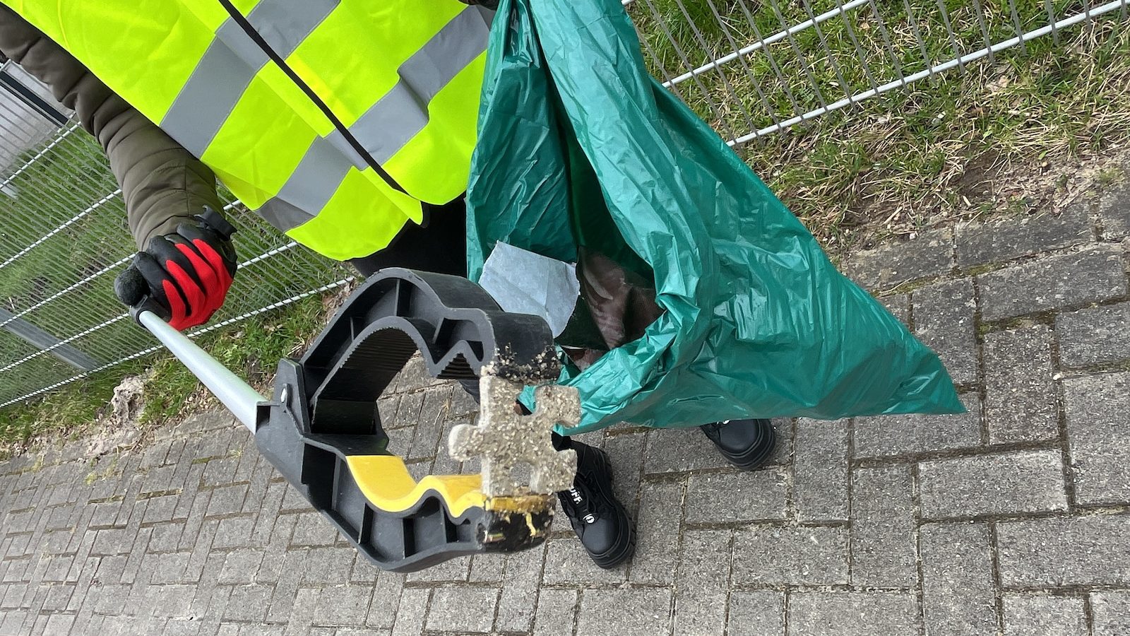 Person in Warnweste sammelt mit einem Greifer ein Bruchstück eines Pflastersteins in einen grünen Müllsack