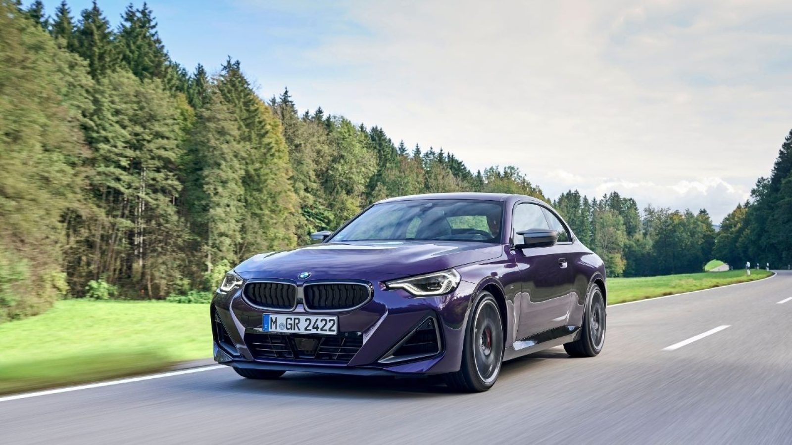 Lila BMW M240i xDrive Coupé auf ländlicher Straße, fährt dynamisch bei sonnigem Wetter an Wäldern vorbei.