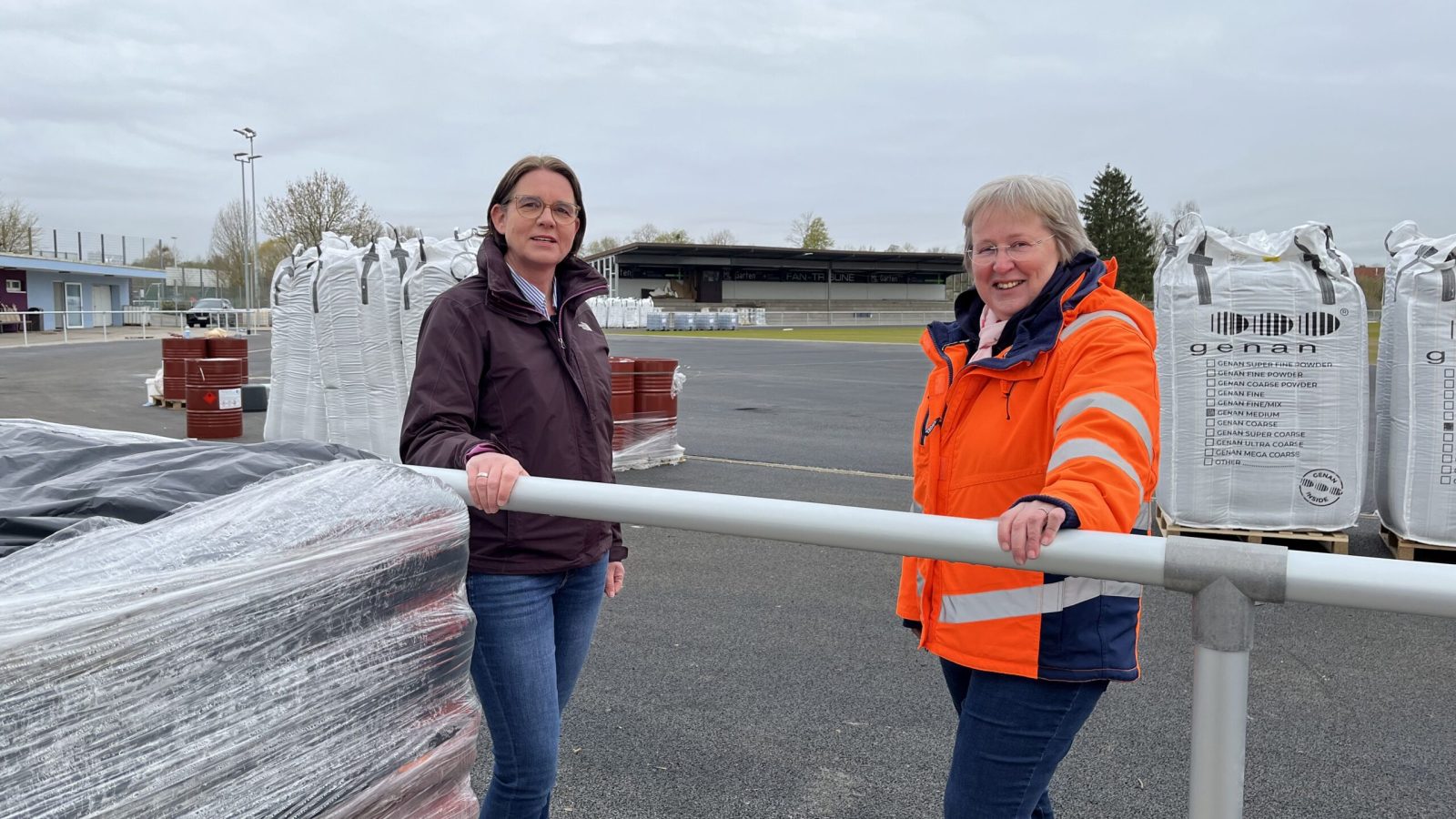 In der Weiling-Arena steht das Material für die Laufbahn-Beschichtung bereit, die Bauarbeiten begleiten (vl.) Karin Hackling (Fachteam Freizeit, Sport) und Renate Rösing (Fachteam FB 70/Tiefbau).