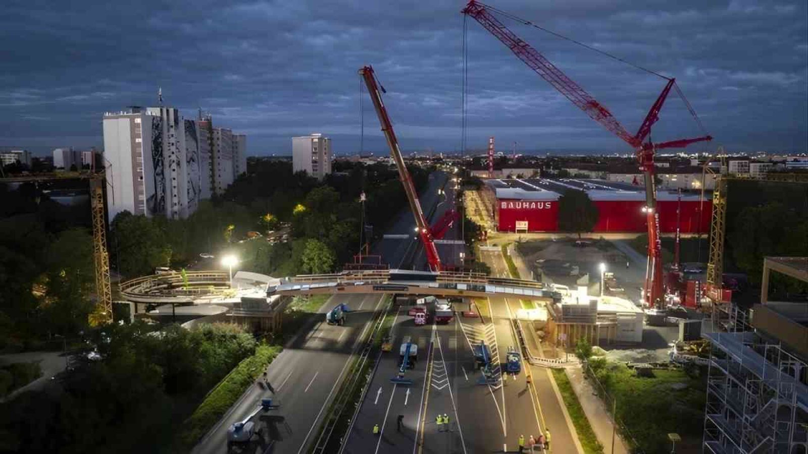 Bauarbeiten an einer Brücke mit Kränen und Bauarbeitern in einer urbanen Umgebung bei Nacht.