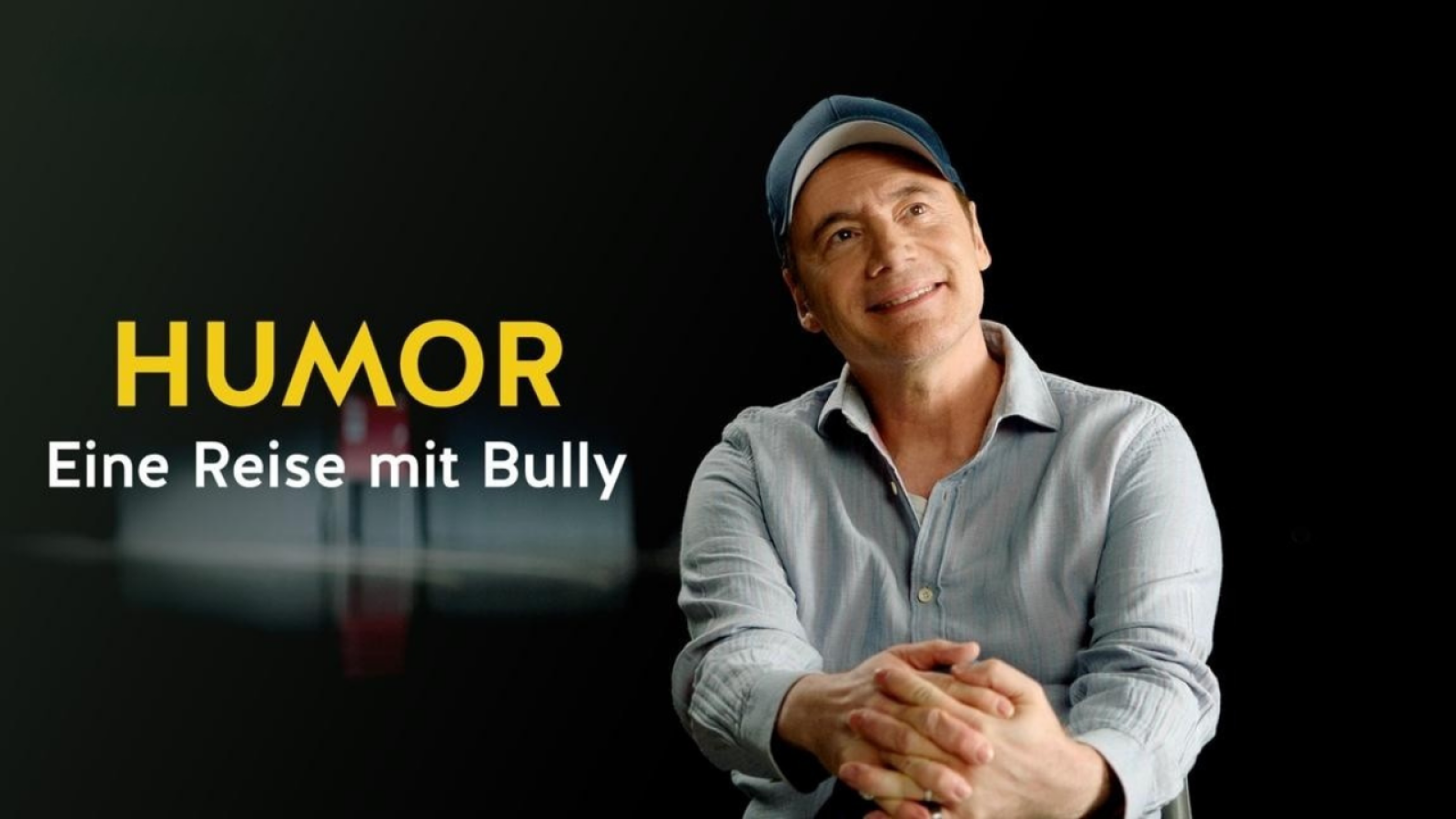 Mann in Hemd und Mütze lächelt, daneben gelber Text Humor - Eine Reise mit Bully auf schwarzem Hintergrund.