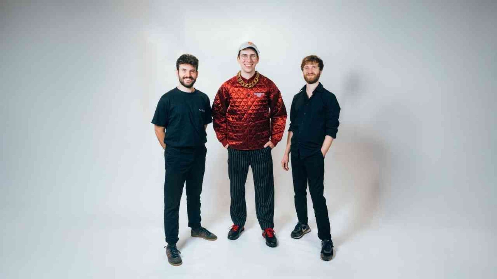 Drei Musiker stehen in lässiger Kleidung vor einem neutralen Hintergrund, bereit für ein Fotoshooting.