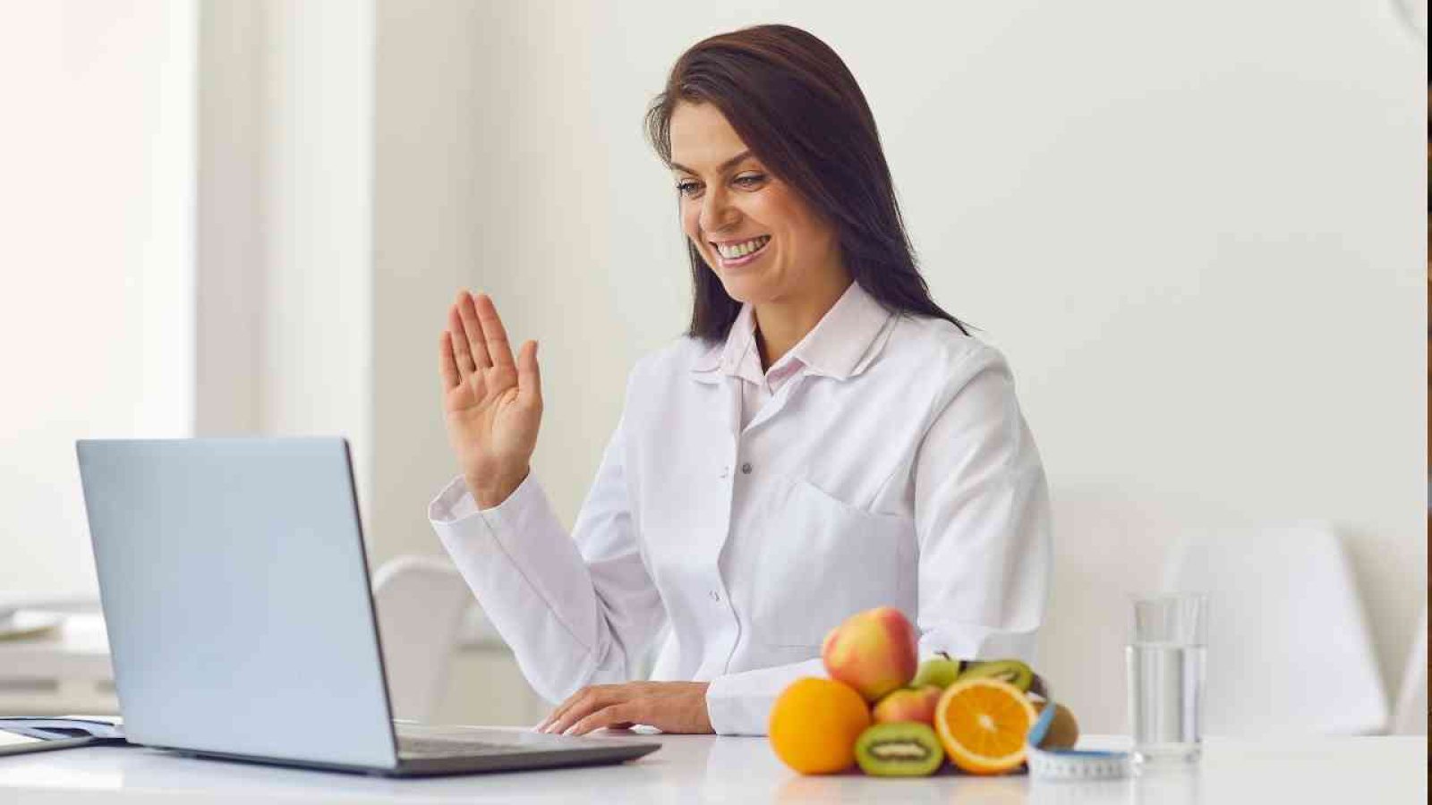 Frau in einem weißen Kittel, die vor einem Laptop sitzt und mit einer Hand winkt, während Obst auf dem Tisch liegt.
