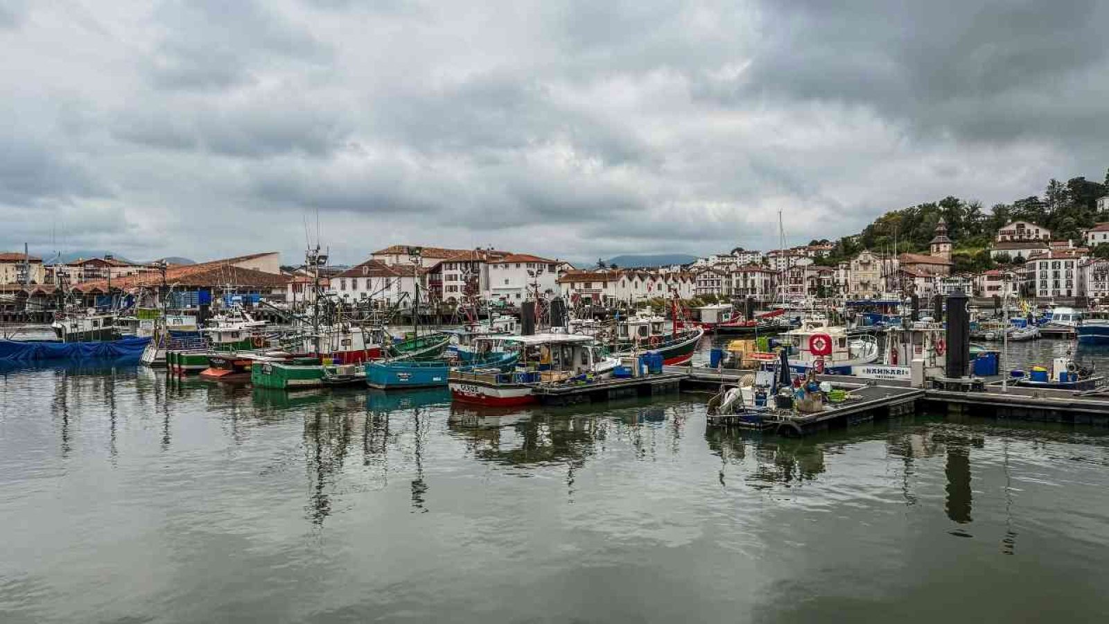 Kleiner Hafen mit bunten Fischerbooten und traditionellen Häusern im Hintergrund unter bewölktem Himmel.