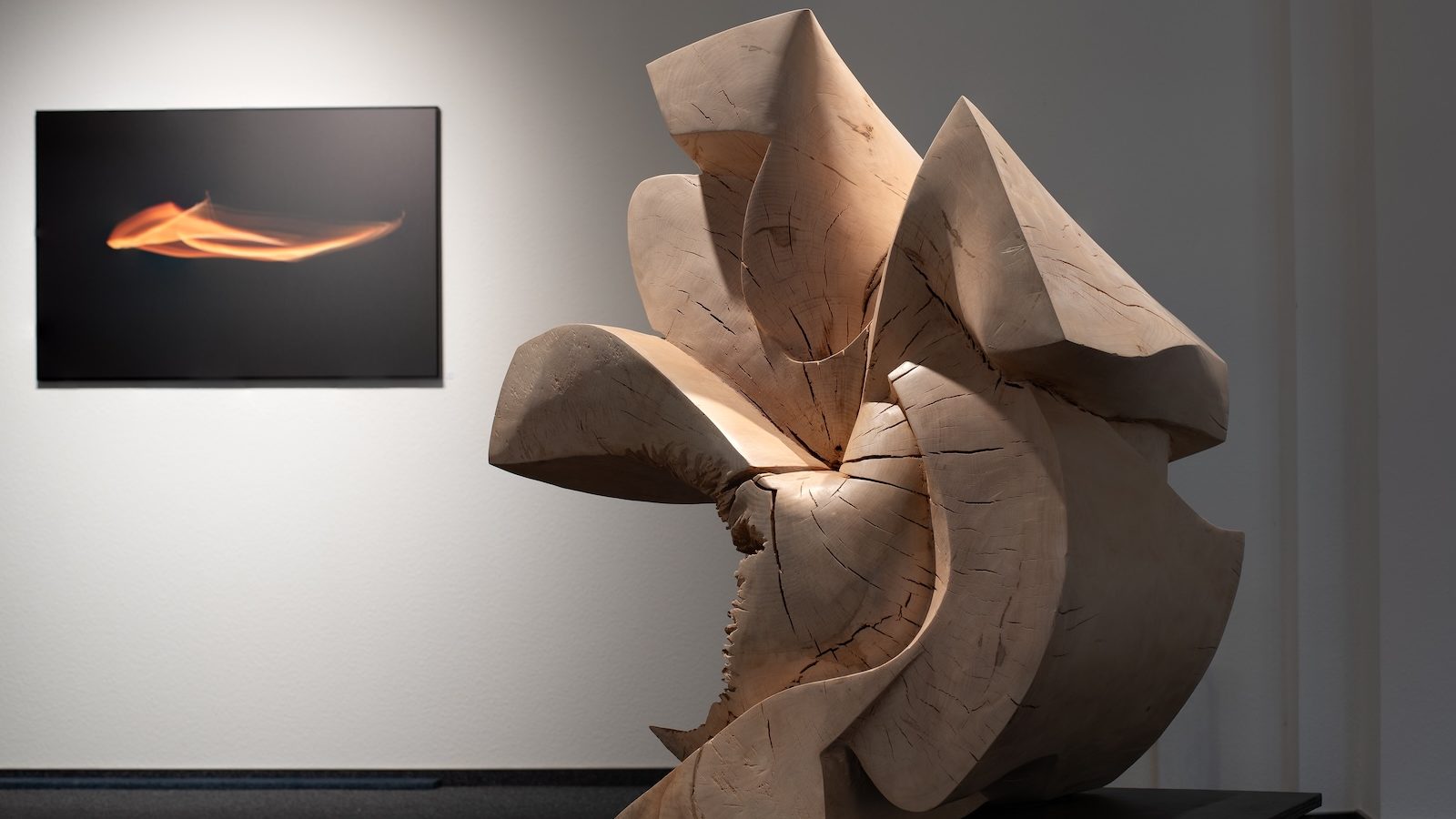 Große Holzskulptur mit geschwungenen Formen vor einer Wand mit einem abstrakten Lichtbild in Orange auf Schwarz