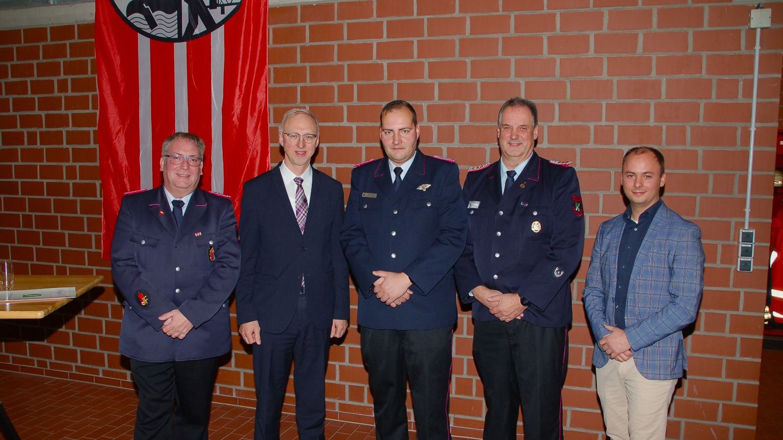 Fünf Männer in Uniformen und Anzügen stehen nebeneinander in einem Innenraum mit einer roten Fahne im Hintergrund.