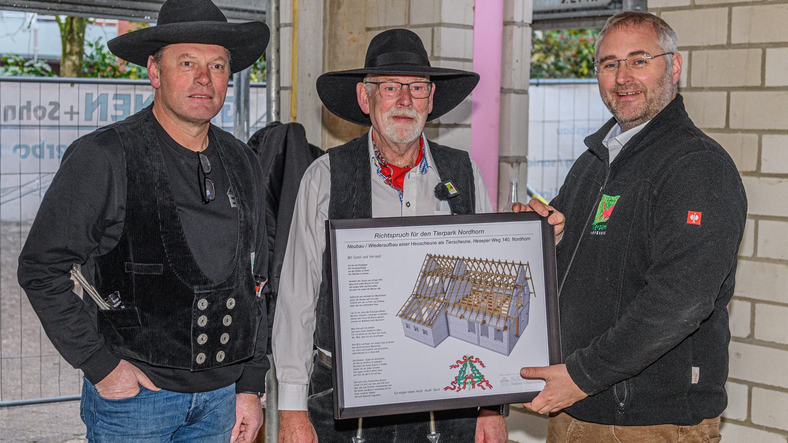 Drei Männer in traditioneller Kleidung präsentieren eine Bauzeichnung eines Holzhauses in einem Innenraum.