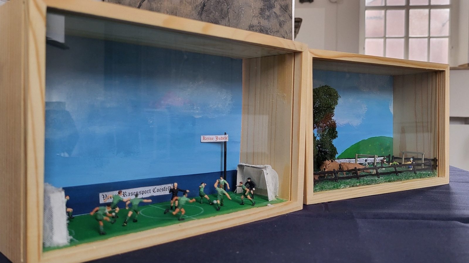 Zwei Schaukästen mit Dioramen, links ein Fußballspiel mit einem Schild Keine Juden, rechts eine Weide mit Kindern hinter einem Zaun