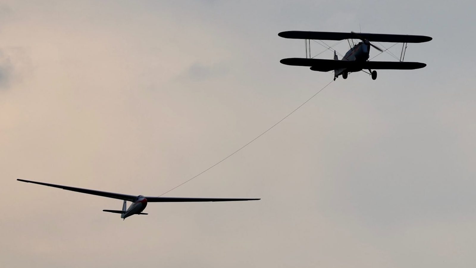 Ein Doppeldecker zieht ein Segelflugzeug an einem Seil in die Höhe, Silhouetten vor grauem Himmel